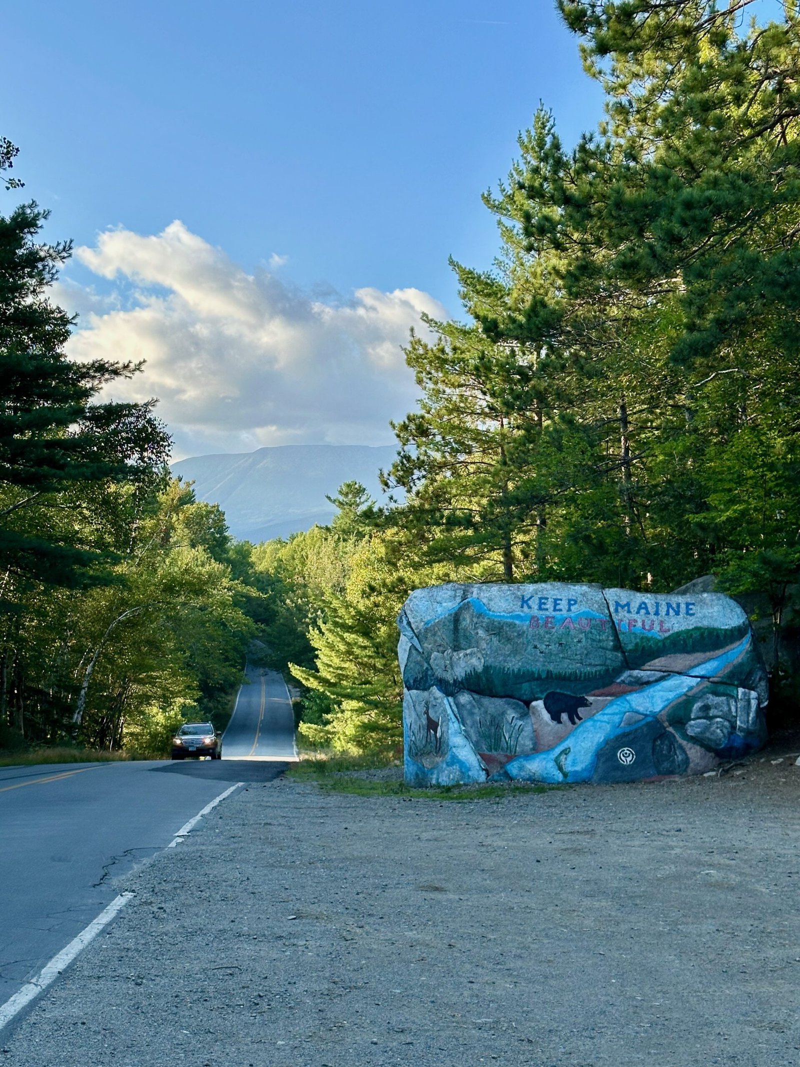 Baxter State Park entrance sign painted on a large rock with a car approaching and Mount Katahdin in the background.