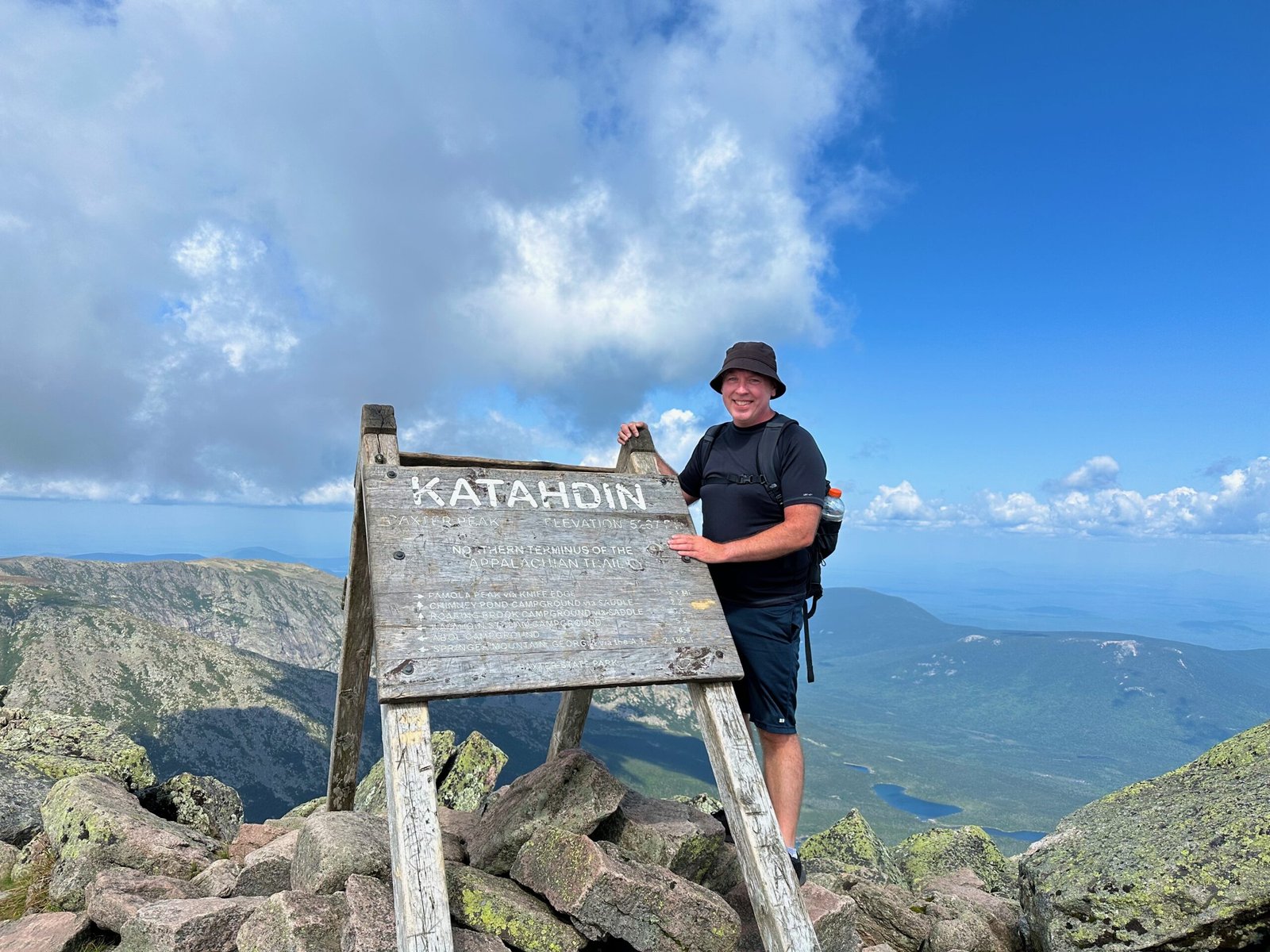 Don Trynor standing beside the Mount Katahdin summit sign with panoramic mountain views, fluffy clouds in the blue sky, and a green carpet of Maine forest in the background.