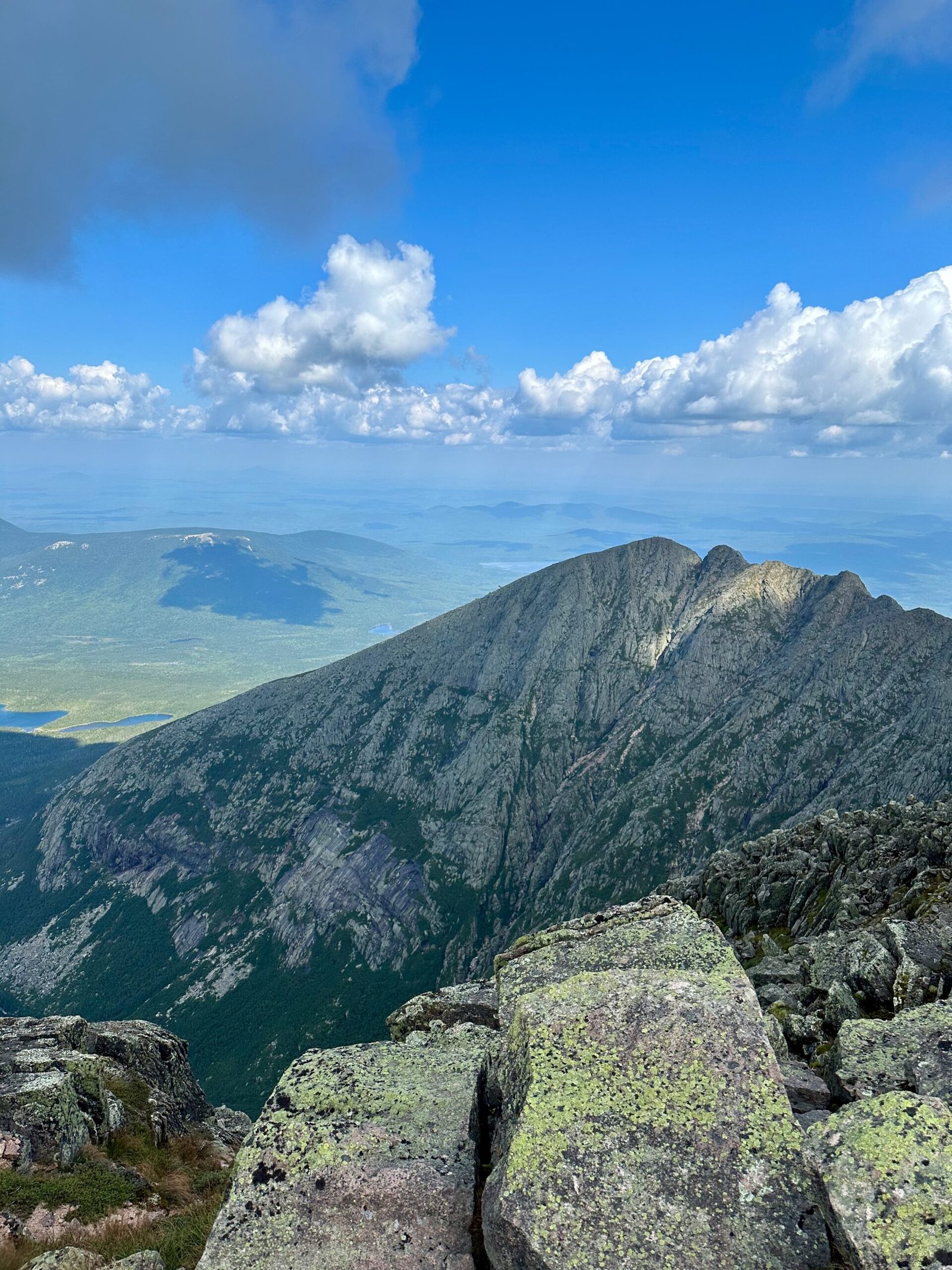 View from the summit of Mount Katahdin showing Pamola Peak and the Knife Edge Trail, with distant peaks of Baxter State Park visible under a clear sky.