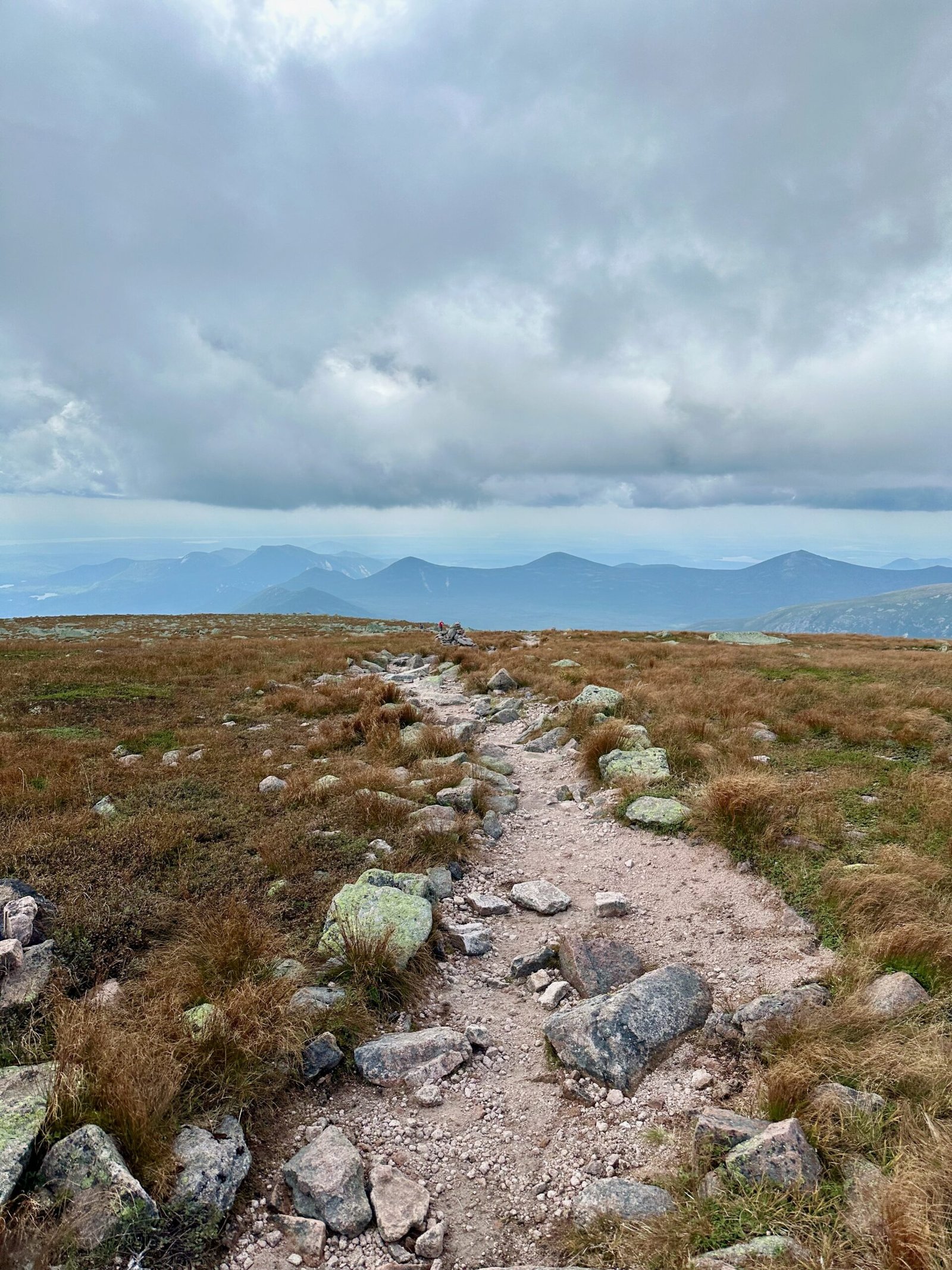 View from the Saddle Trail near the summit of Mount Katahdin, showing the trail through ecologically sensitive alpine plants with distant peaks of Baxter State Park in the background.