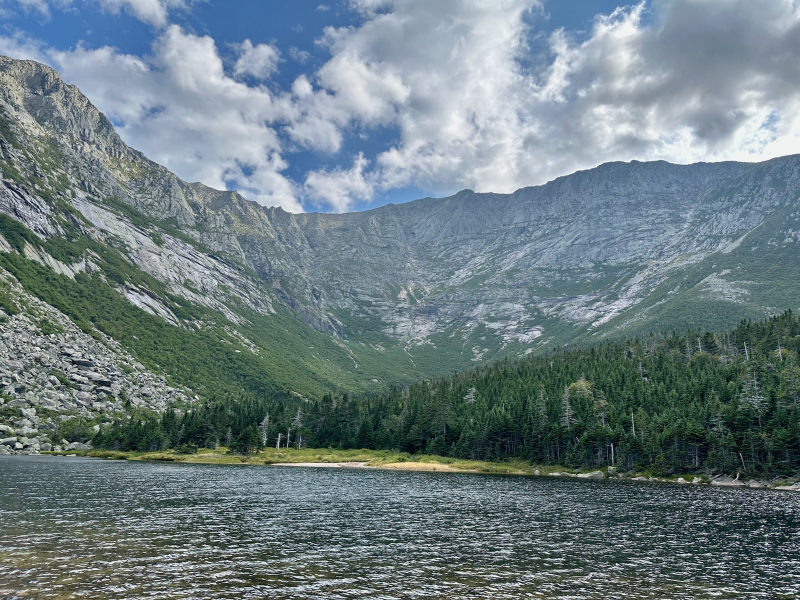 View from the shore of Chimney Pond with the pond’s water in the foreground, forest, and talus slopes rising up Mount Katahdin in the background.