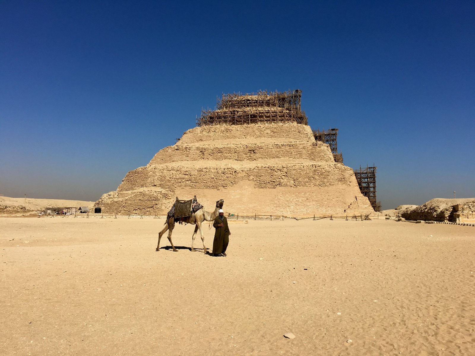 Pyramid of Djoser with a man walking beside a camel, wooden staging partially covering the structure.