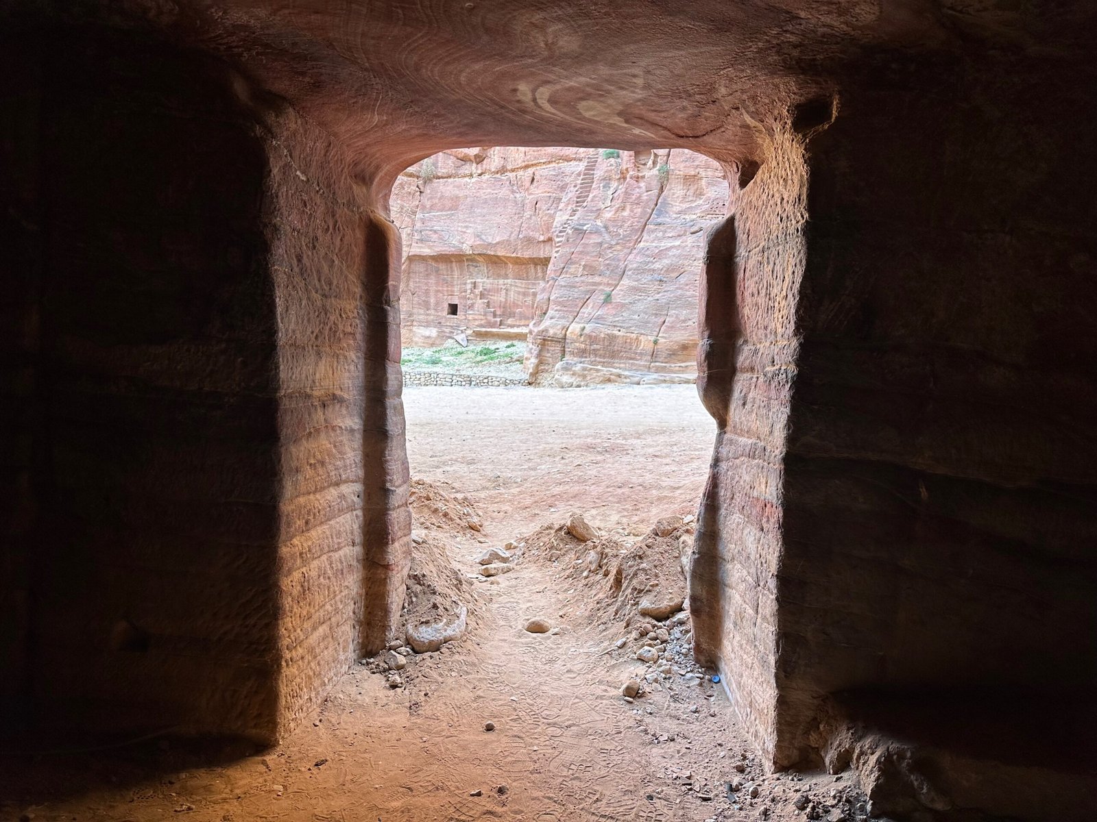 View from inside a Petra cave dwelling, looking out toward the sandy canyon floor and the sandstone canyon wall on the opposite side.