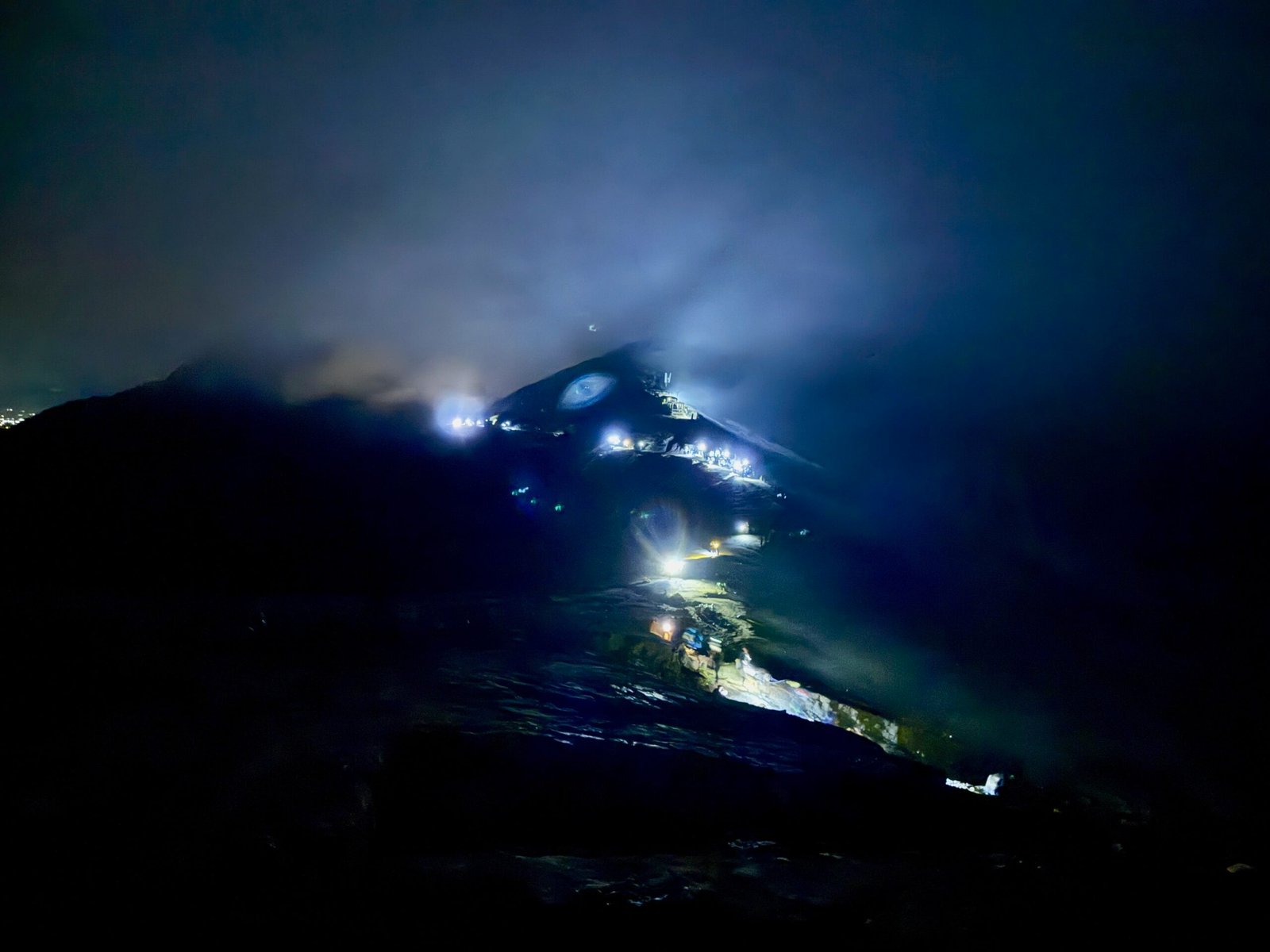 Hikers wearing headlamps trek along the steep trail to Ijen Volcano’s caldera in Indonesia, forming an eerie line of light in the cloudy darkness.