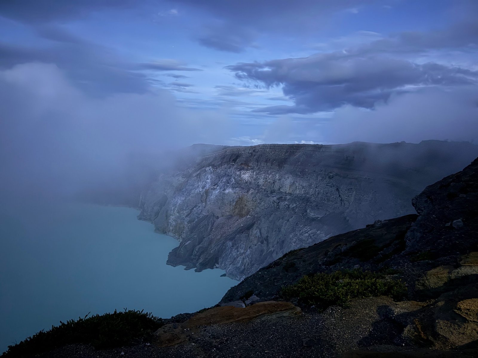 A wide view of Ijen Volcano’s acidic turquoise lake at dawn, seen from the caldera rim against a pale blue sky in East Java, Indonesia.