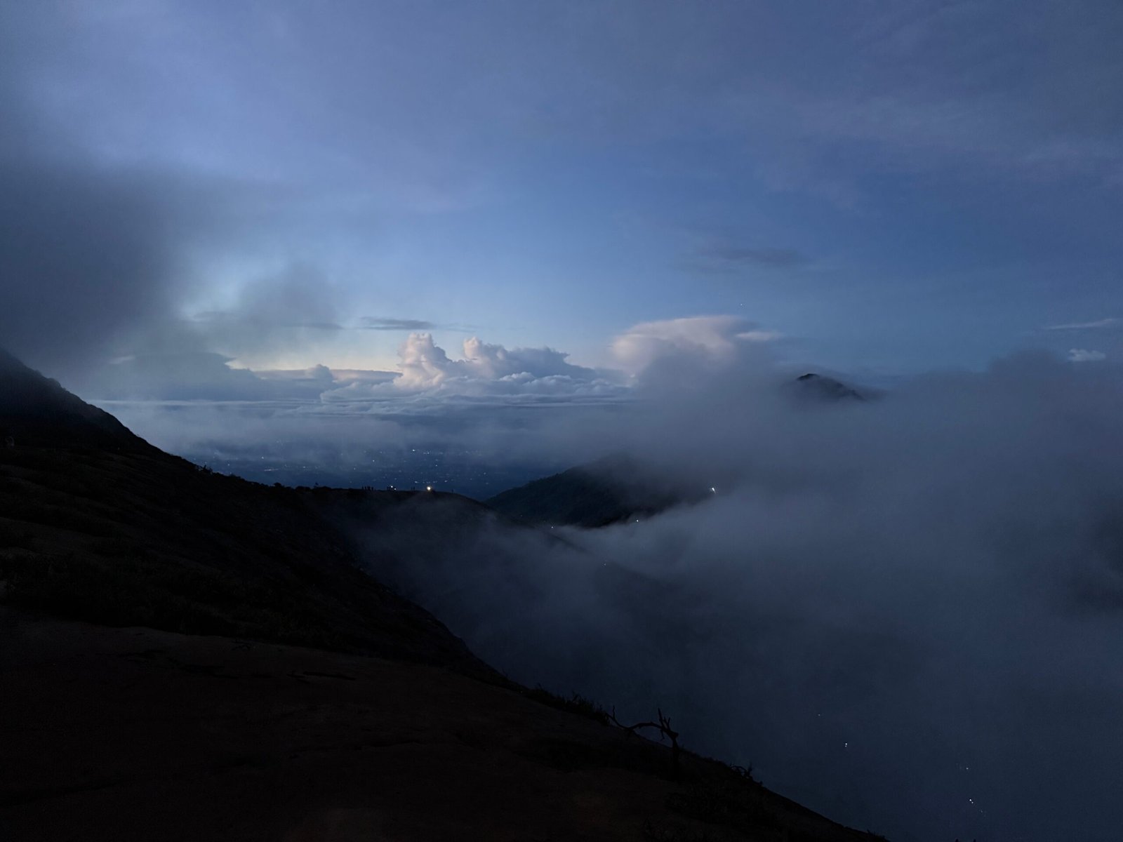 A panorama of Banyuwangi at sunrise from the Ijen Volcano summit, with blue-hued skies and billowing clouds rising over the ocean.