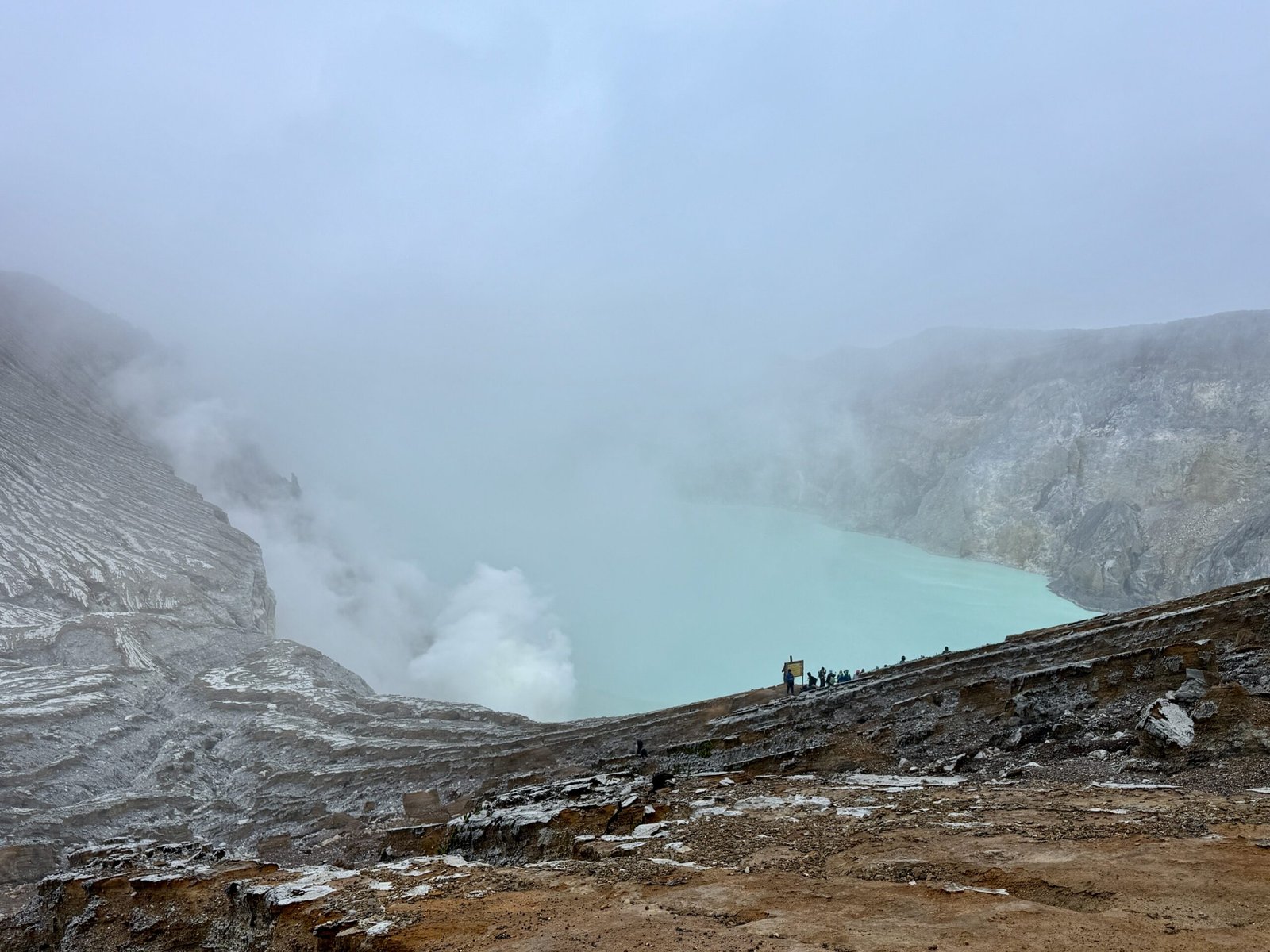 Daylight overhead view of Ijen Volcano’s turquoise acidic crater lake with plumes of sulfuric gases rising from vents below.