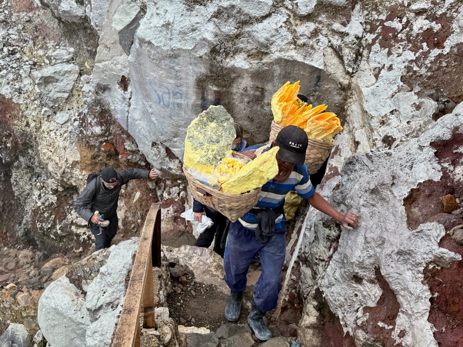 An Ijen Volcano sulfur miner climbs toward the caldera rim carrying a heavy basket of yellow sulfur rock without wearing protective gear.