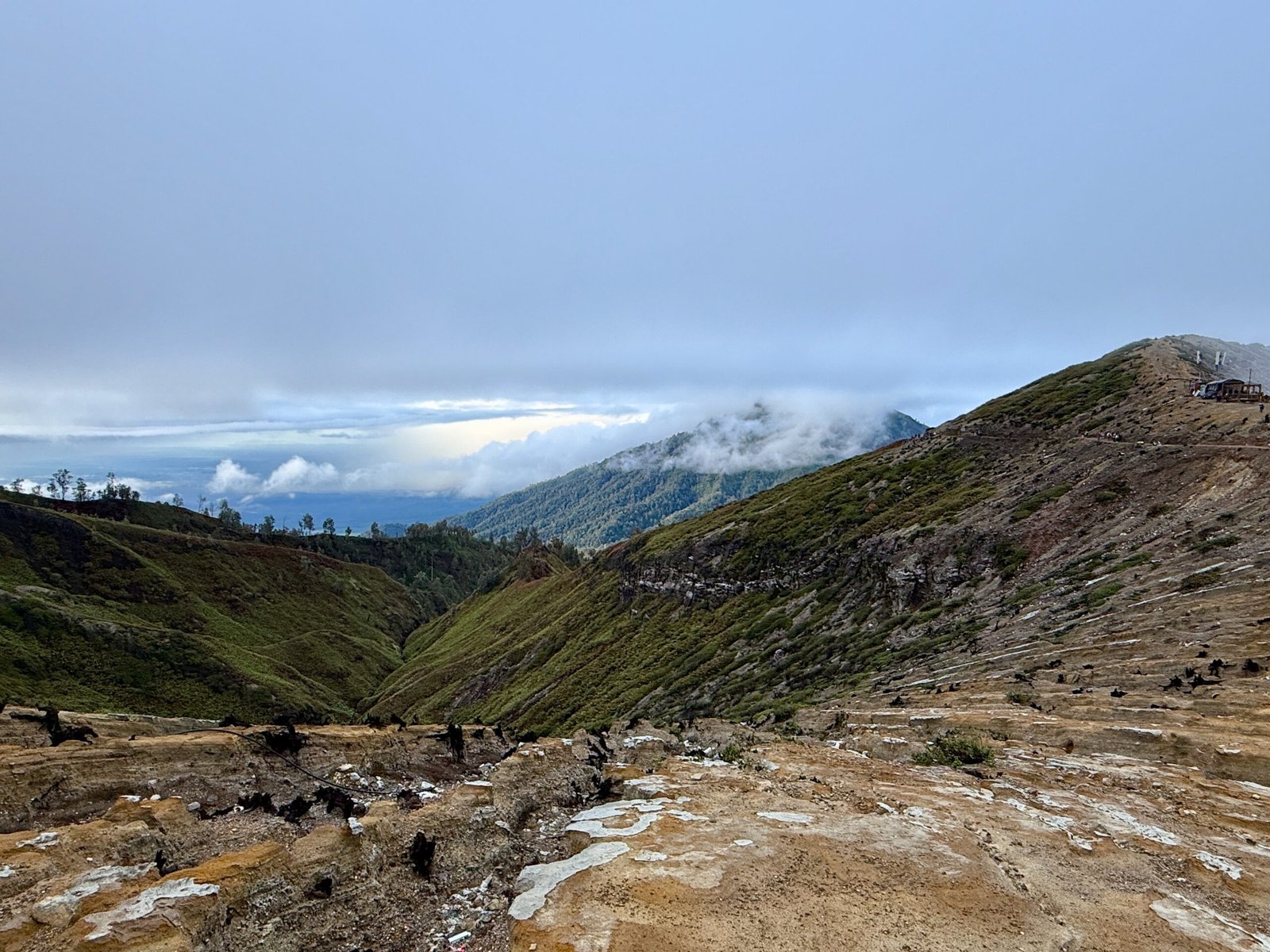 A daytime view from the Ijen Volcano rim showing green, cloud-shrouded slopes and forested terrain facing Banyuwangi.