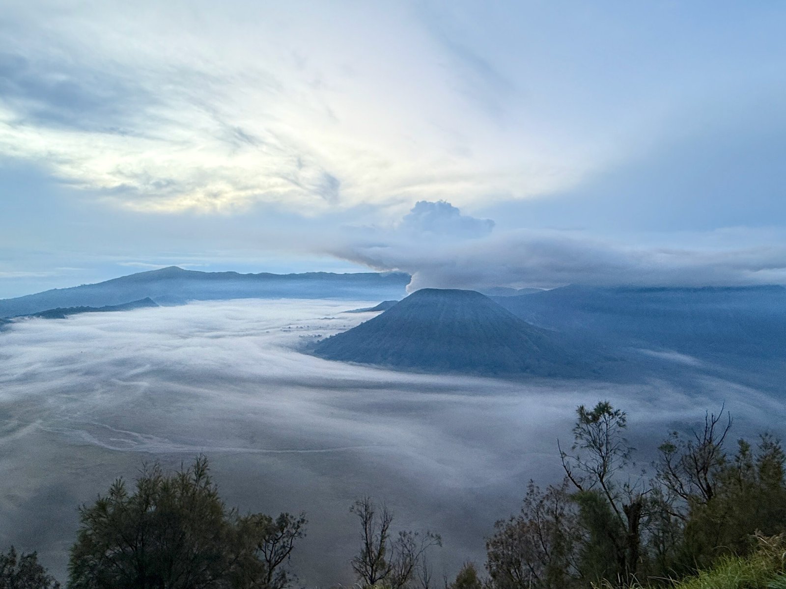 Panoramic view of Mount Bromo and Mount Batok at sunrise on a cloudy morning, with smoke rising from Mount Bromo’s active caldera.