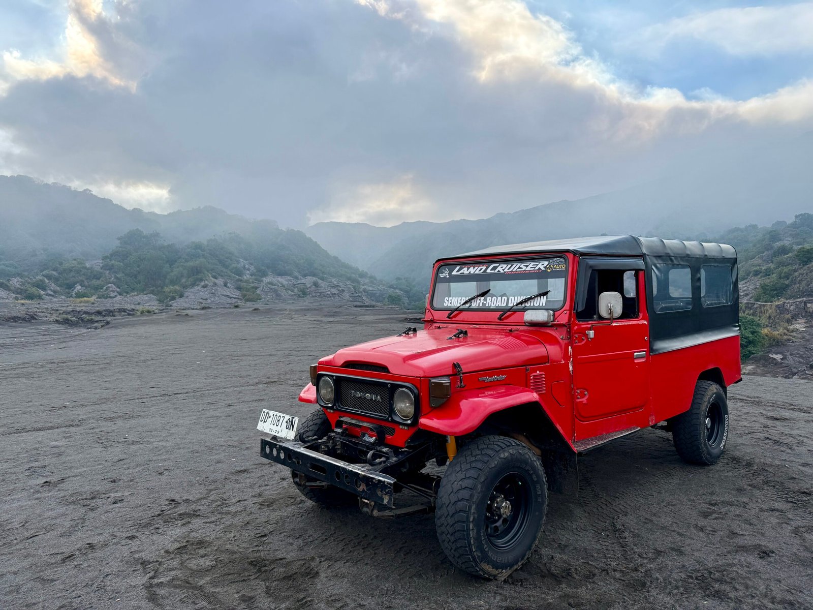 Tour jeep in the foreground with Mount Bromo’s steaming caldera in the background, set in the black volcanic ash plains of the Tengger Caldera.