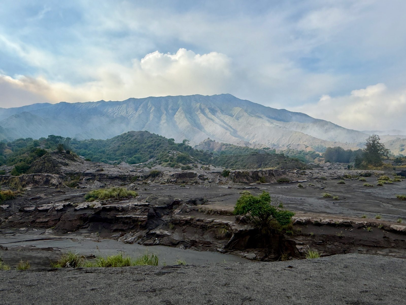 Eroded volcanic landscape in the Tengger Caldera, covered in black ash, with the caldera’s high rim visible in the background.