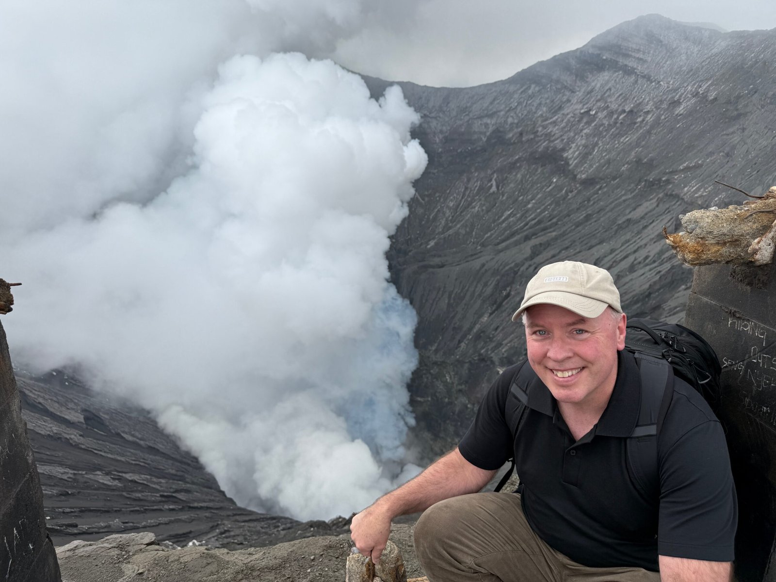 Don Trynor kneeling on the rim of Mount Bromo’s caldera, with smoke and steam rising dramatically from the volcano.