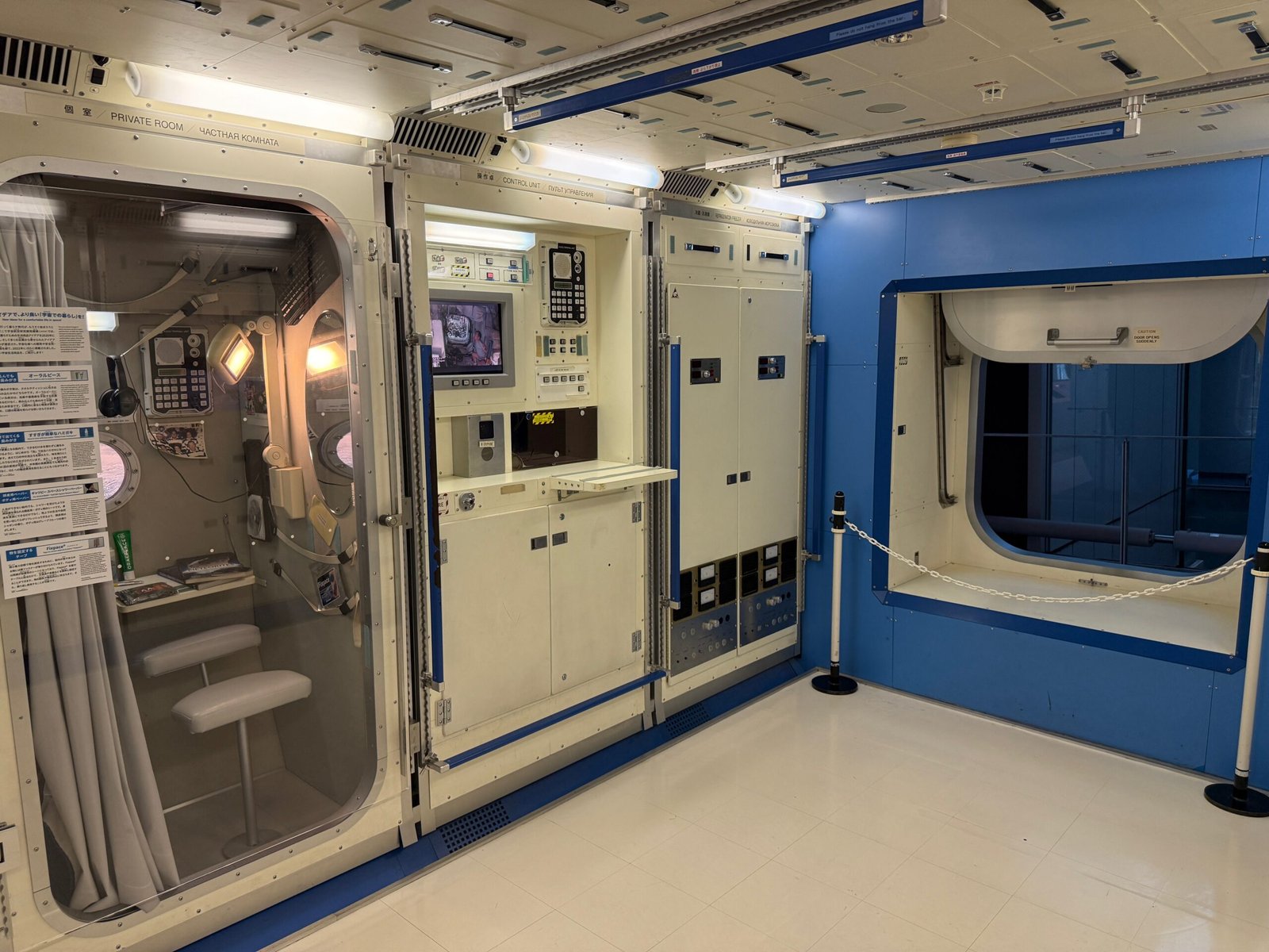 Interior view of the ISS replica at Miraikan, showing a private room, control panel, and refrigerator/freezer unit.