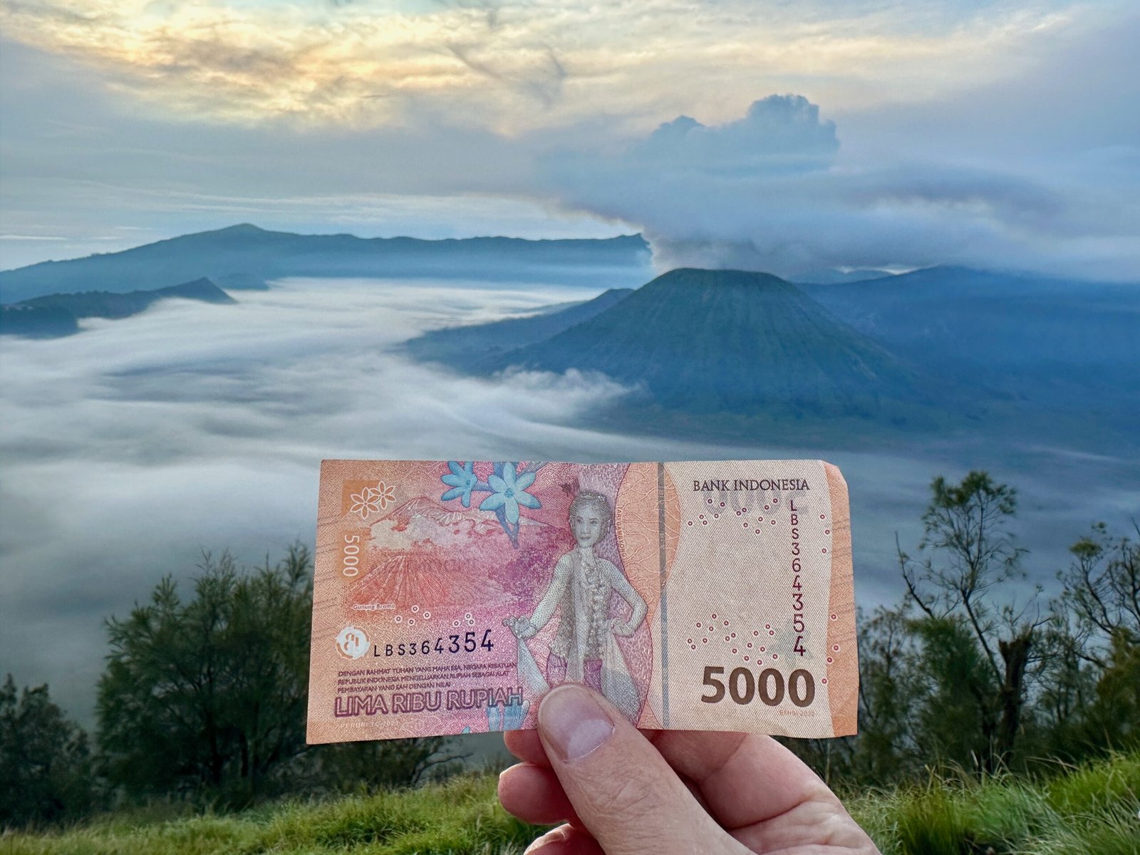 5,000 Indonesian Rupiah note depicting Mount Bromo, held up with the actual Mount Bromo and its sunrise view in the background.