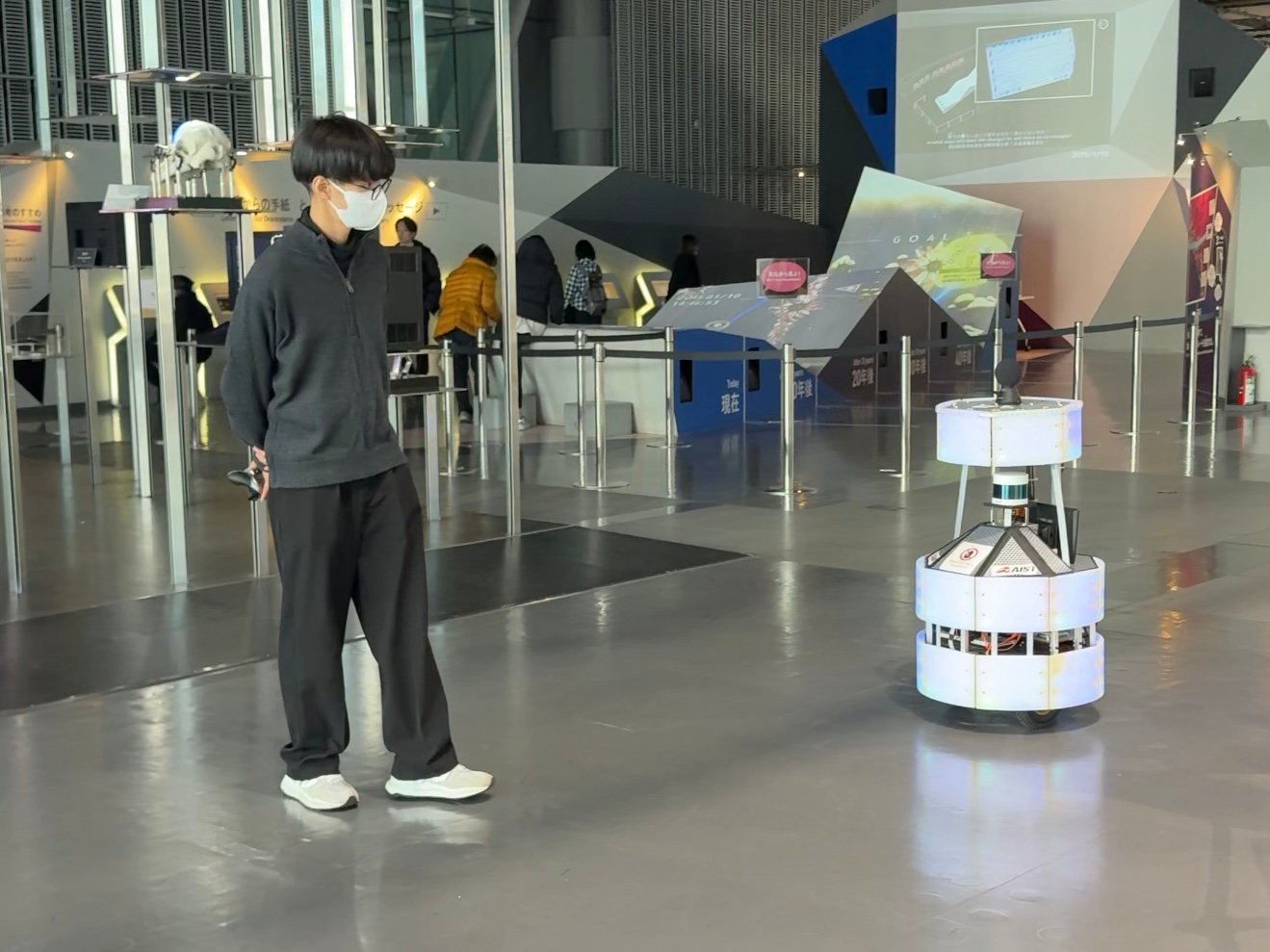 Prototype robot moving around the robotics exhibit at Miraikan, followed closely by a human assistant.