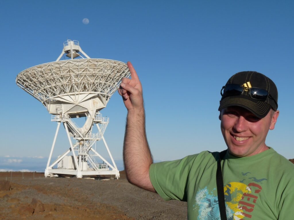 Don Trynor standing in front of a radio telescope at the summit of Mauna Kea, pointing at the sky.