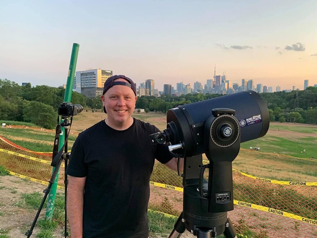 Don Trynor next to an astronomical telescope at dusk in Riverdale Park, Toronto, with the downtown skyline in the background, preparing to observe the Neowise comet.