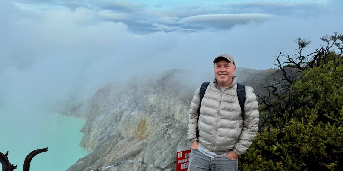 Don Trynor, author at Curious Don, stands on the rim of the Ijen Volcano caldera in Indonesia, with a turquoise crater lake visible below.