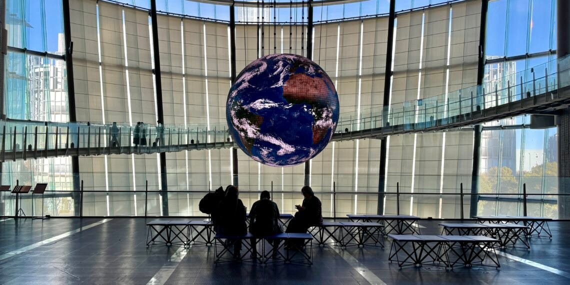 Silhouetted visitors sitting at a table in front of the illuminated Geo-Cosmos globe in Miraikan’s main hall.