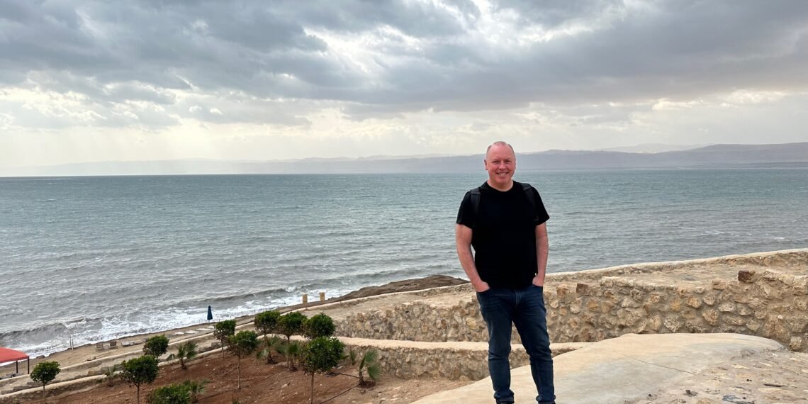 Don Trynor, author of the Curious Don website, standing in front of the Dead Sea in Jordan, with the west bank of the Dead Sea in Israel in the background.